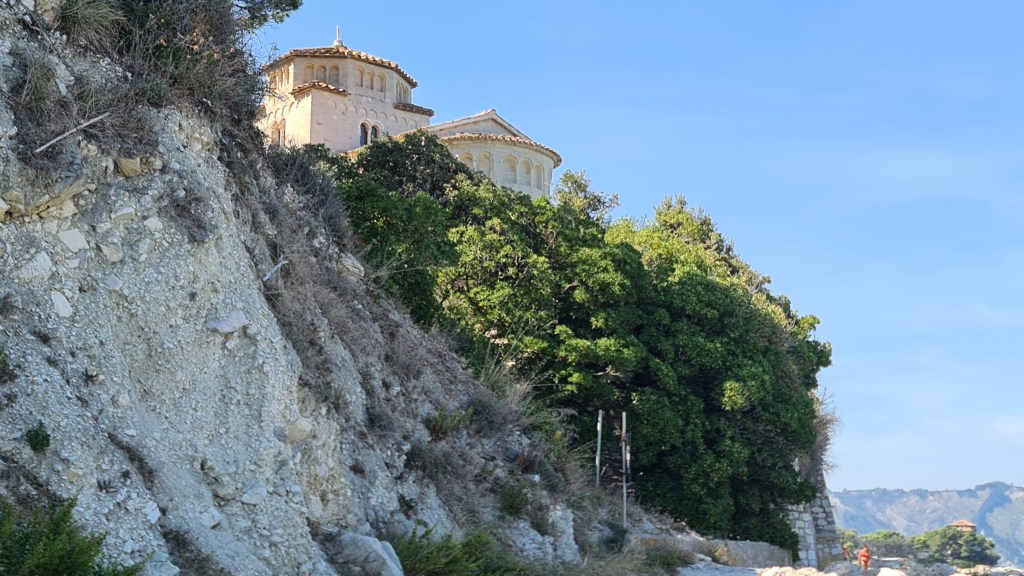 A hillside view featuring a Mediterranean-style building surrounded by dense green foliage and rocky terrain.