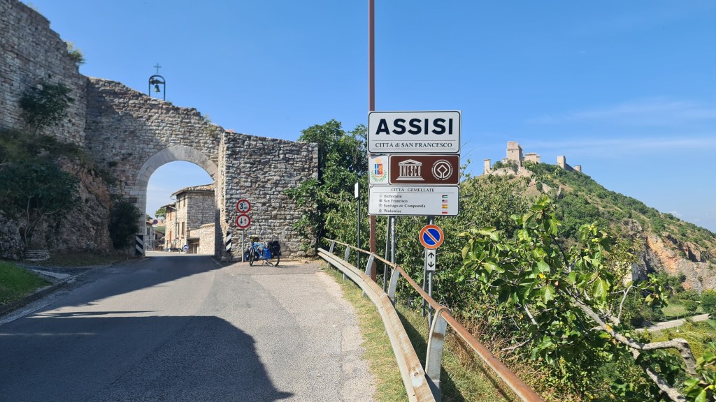 View of the entrance to Assisi, Italy, with a stone archway, road signs, and a hillside castle in the background.