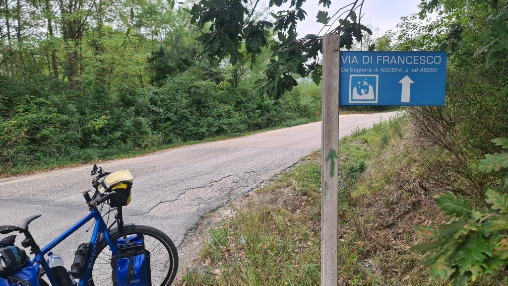 A blue sign indicating 'Via di Francesco' with directions, located on a rural road surrounded by greenery, with a bicycle leaning against a post.