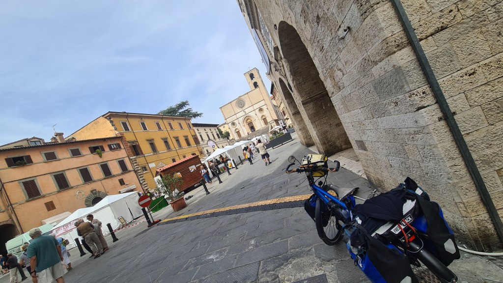 Een fietser staat met een blauwe fiets en bagage in een pittoreske straat in Todi, Italië, met uitzicht op een historische kerk en bezoekers van een markt.