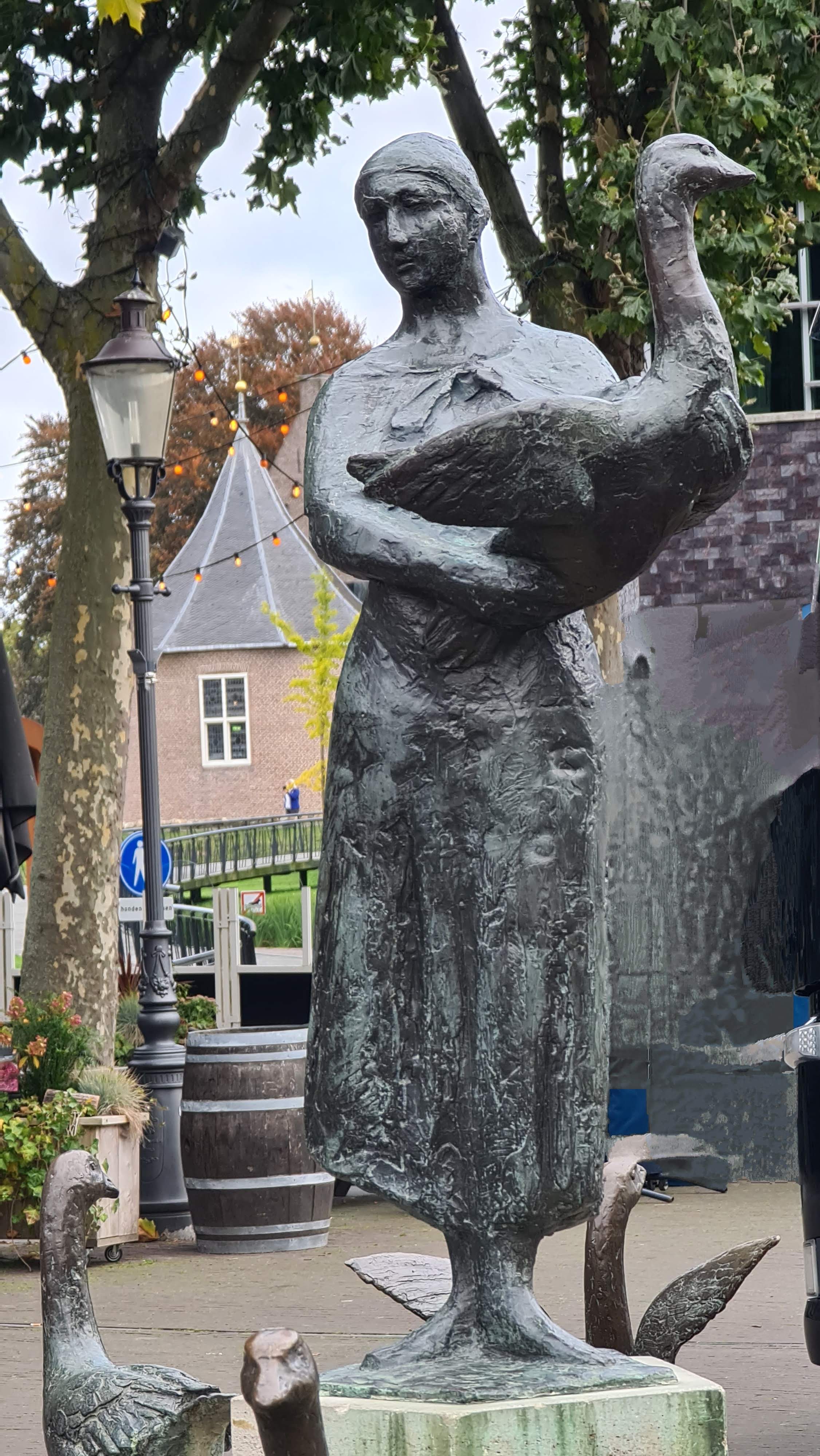 A bronze statue of a woman holding a goose, surrounded by smaller goose sculptures in an outdoor setting.