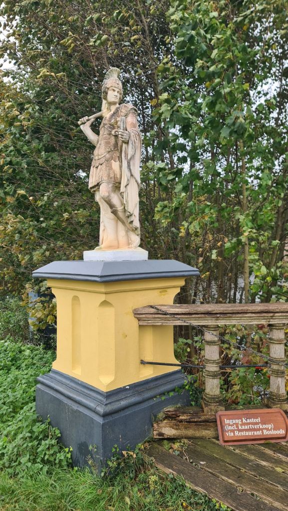 A sculpture of a Roman soldier stands on a yellow pedestal, surrounded by green trees. A wooden sign nearby indicates the entrance to a castle.