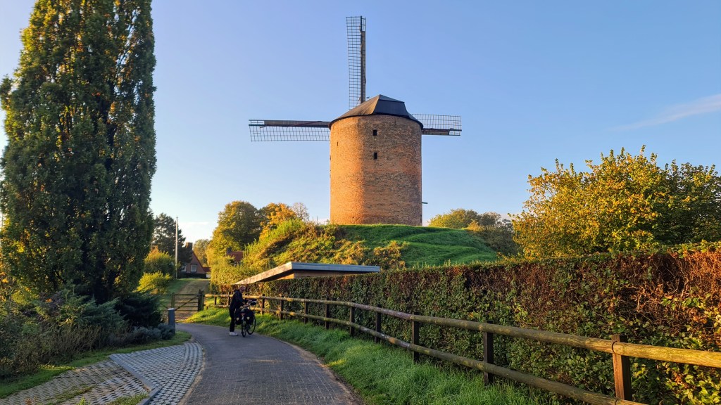 A scenic view of a historic windmill on a grassy hill, surrounded by trees and a paved path where a person is riding a bicycle.