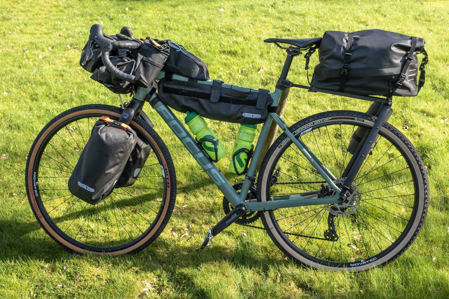 Gravelbike of hybride fiets? | Welke past bij jouw fietsvakantieGray touring bicycle loaded with multiple black waterproof pannier bags and two green water bottles on a grassy field