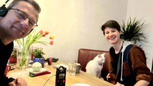 A man wearing headphones and a woman smiling at a table with a white cat and a recording device, surrounded by a vase of tulips and glasses of water.