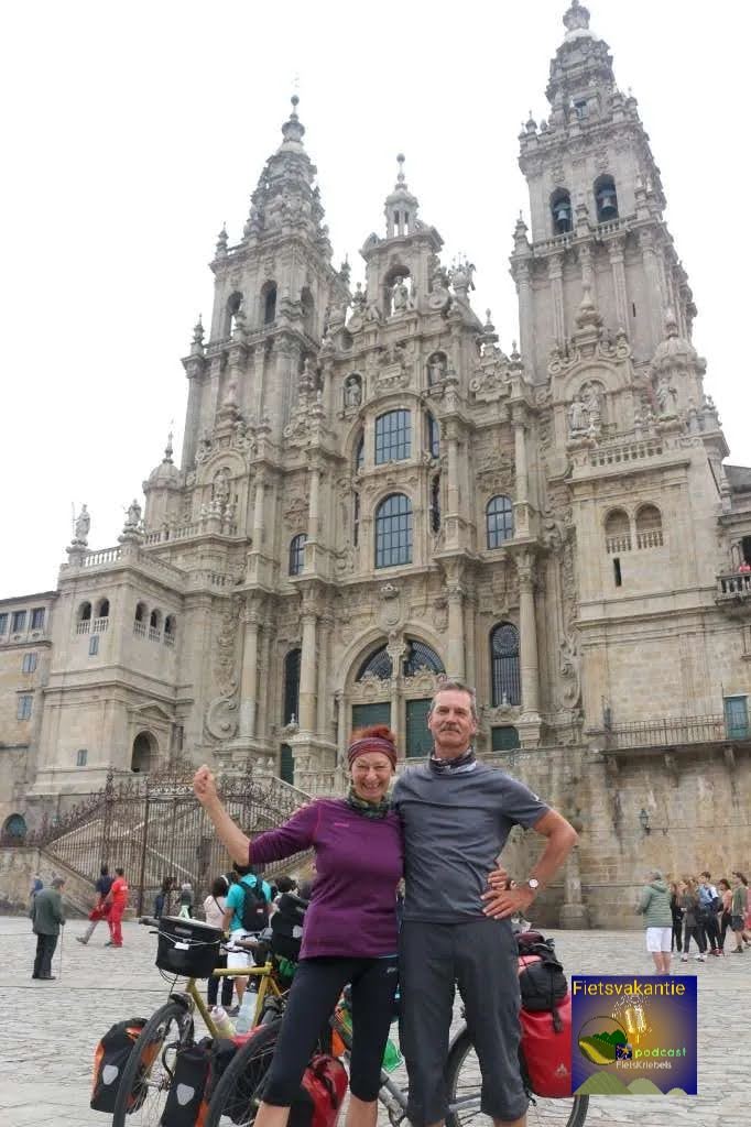 A couple stands in front of an ornate building, smiling and posing with their bicycles. The structure features intricate architectural details with multiple spires.