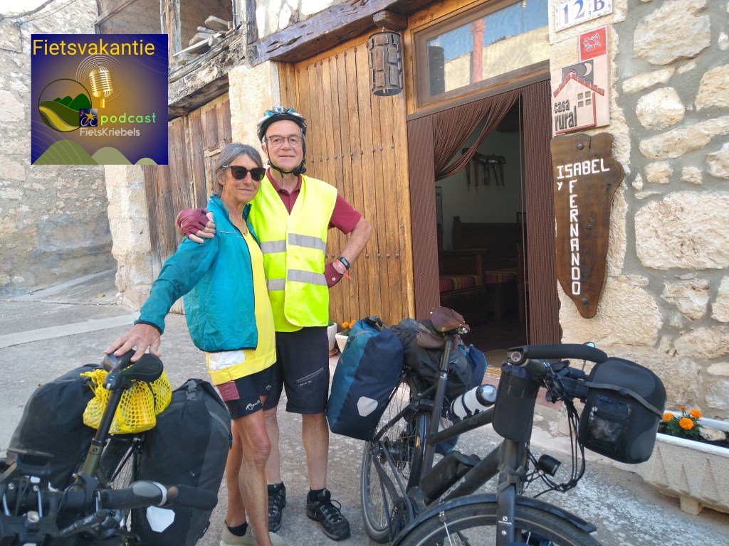 A couple poses in front of a stone wall building with a wooden door, each wearing cycling gear and surrounded by bicycles with panniers. The scene includes a podcast logo in the corner.