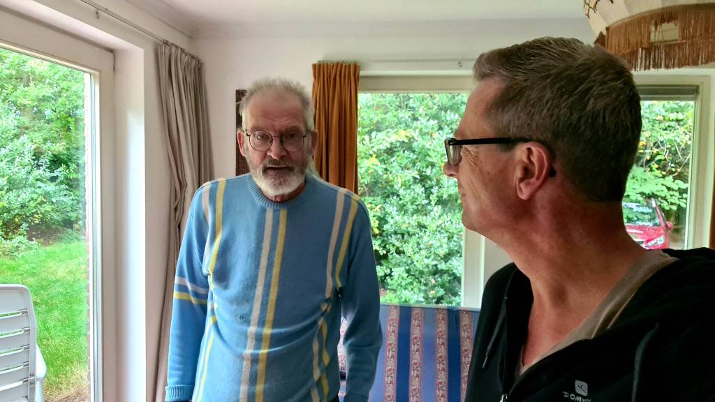 An older man with glasses and a beard wearing a blue sweater stands inside a room looking at a younger man with glasses. There are plants visible outside through a window.