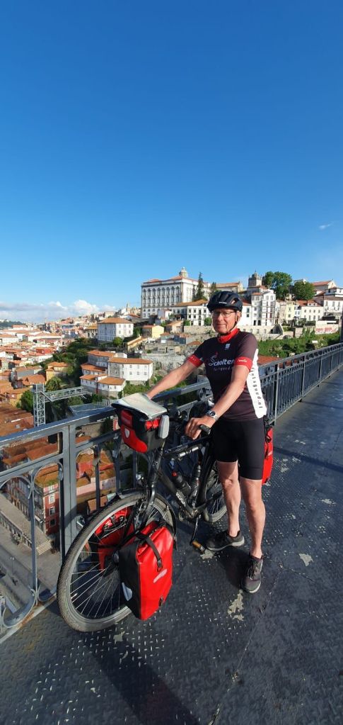 Fietser met helm staat naast een fiets met bagage, op een uitzichtpunt over de stad Coimbra onder een blauwe lucht.