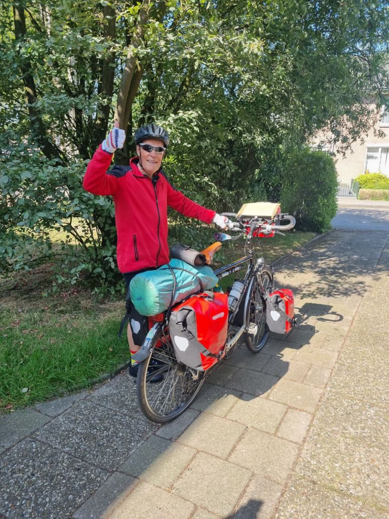 Een enthousiaste oudere fietser in een rode jas en fietshelm terwijl hij zijn hand opsteekt voor de camera, naast een goed beladen touringfiets.