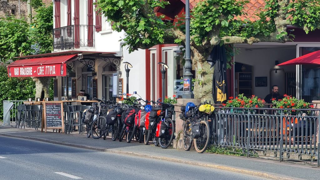 Een café met een rode luifel en een terras, omringd door bomen, met fietsers en fietsen in de voorkant.