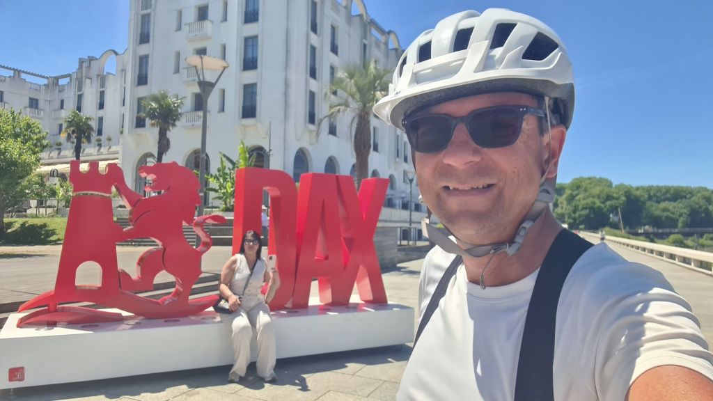 Een man in een fietshelm en zonnebril staat glimlachend voor een groot, rood DAX-beeld in een zonnige omgeving, met een vrouw op de achtergrond die zit en een smartphone vasthoudt.