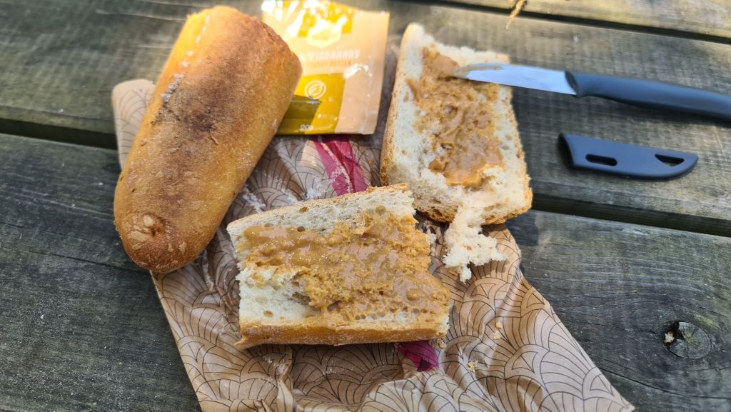Een gesmeerd broodje met pindakaas op een houten tafel, naast een broodje en een mes, met een verpakking van energierepen op de achtergrond.