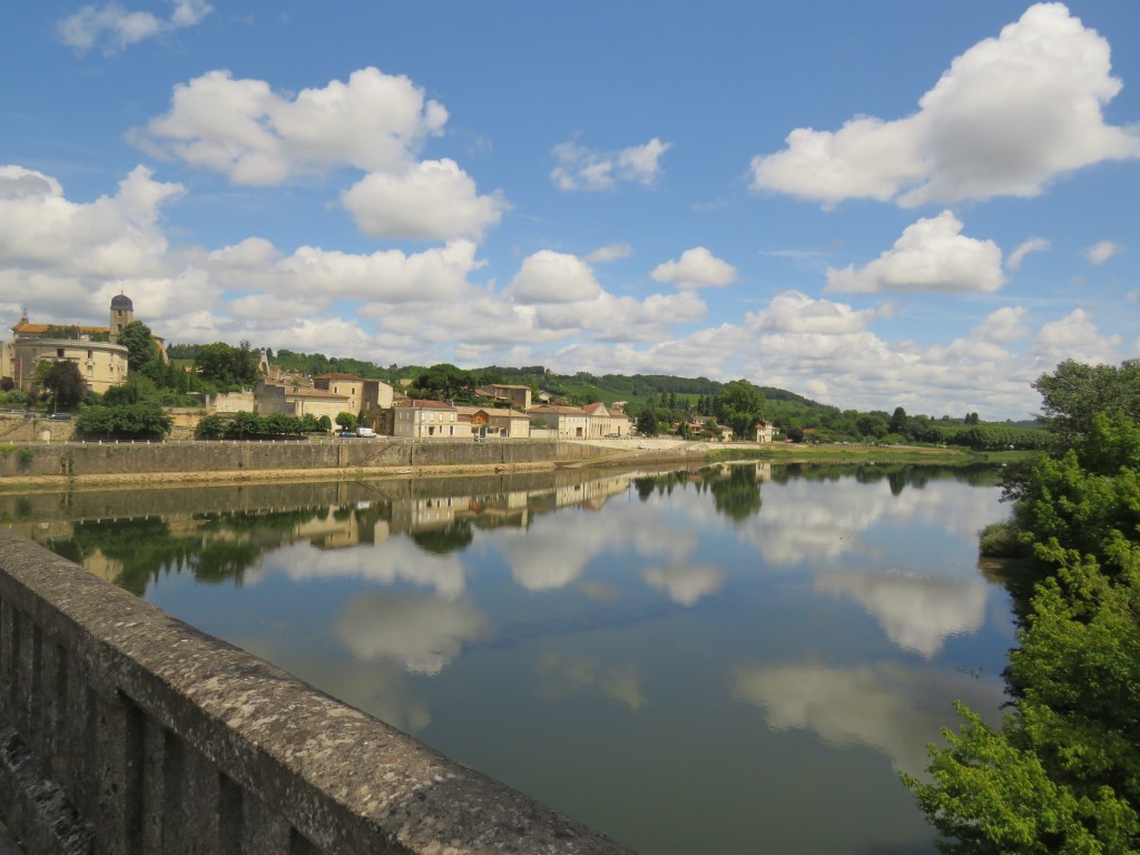 A scenic view of a calm river reflecting the sky and clouds, with a quaint village along the riverbank surrounded by green hills.