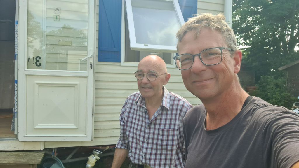 Een selfie van twee mannen buiten een stacaravan, met een open deur en een raam op de achtergrond.