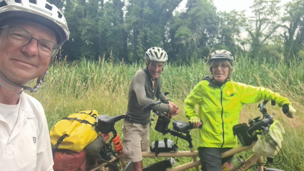 Groep van drie fietsertjes op een tandem met kleurrijke fietstassen, staande op een groene achtergrond met gras en bomen.