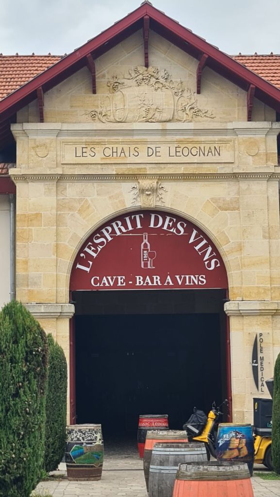 Uitzicht op de ingang van L'Esprit des Vins, een wijnbar en -cave, met houten vaten aan de zijkanten en een stenen gevel met inscripties.