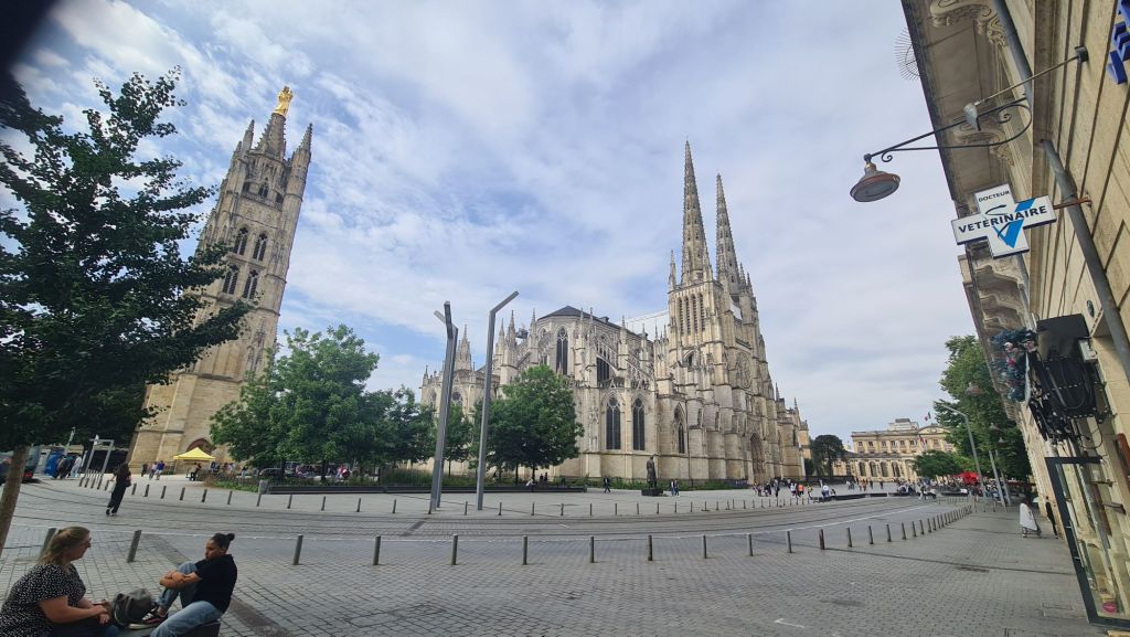 Zicht op de indrukwekkende kathedraal Saint-André in Bordeaux met zijn hoge torenspitsen, omringd door groen en een plein met mensen.