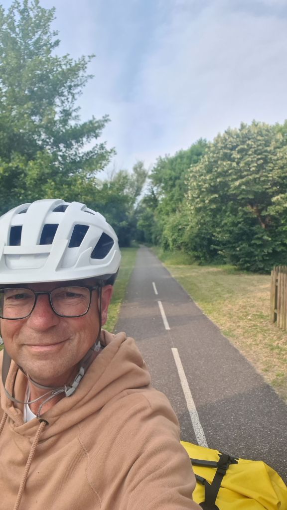 Een glimlachende fietser in een helm, op een fietspad omringd door groene bomen en begroeiing.