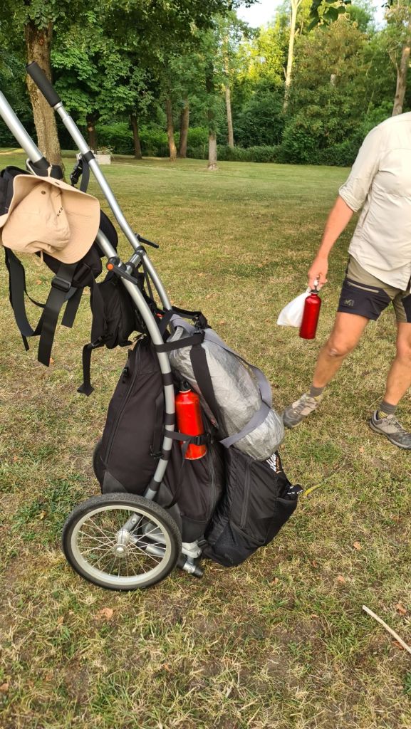A walking cart with a water bottle attached, positioned on grass, outdoors. A person is seen holding a red water bottle and wearing a light-colored shirt.