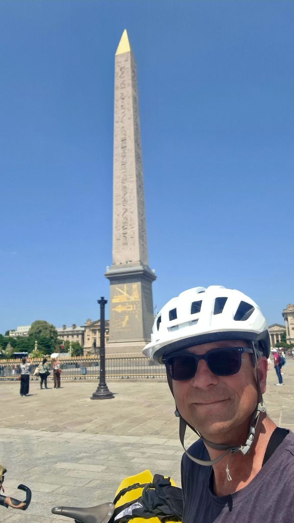 Een fietser poseert voor een grote obelisk onder een heldere blauwe lucht in Parijs.