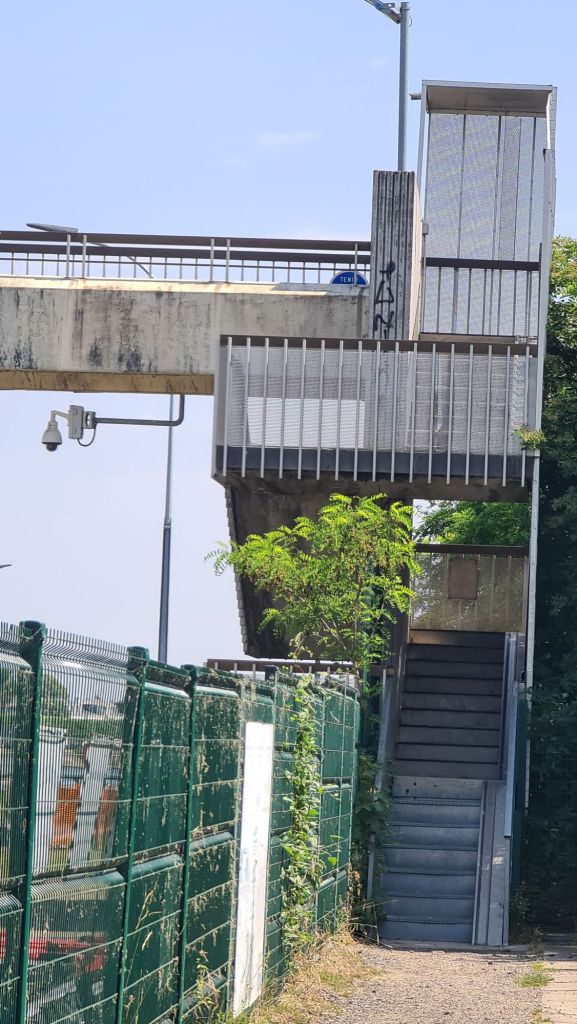 Een trap naar een verhoogd pad of overpass, met hekwerk aan beide zijden en omringende vegetatie.