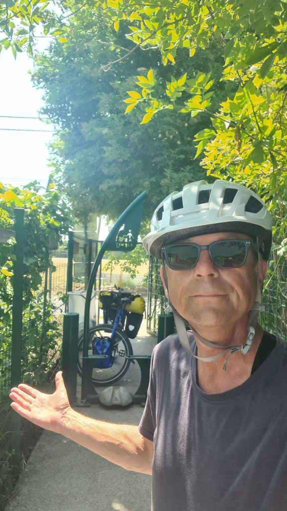 Een fietser in een helm en zonnebril staat buiten, met een fiets met bagage achter zich, omringd door groene bladeren en een hek.
