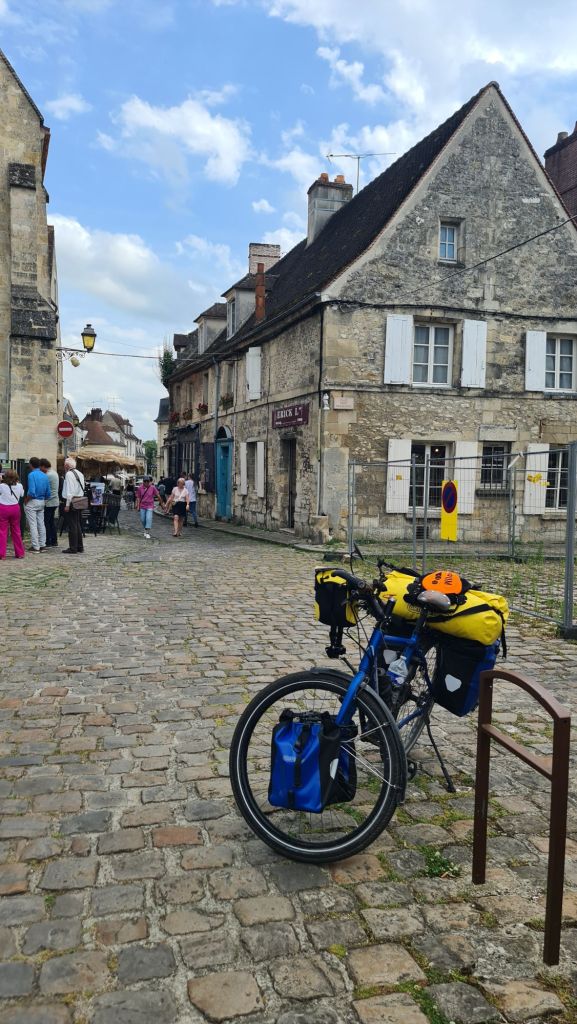 Een blauwe fiets met fietstassen staat geparkeerd op een geplaveide straat in het middeleeuwse stadje Senlis, omringd door oude stenen gebouwen en enkele wandelaars.