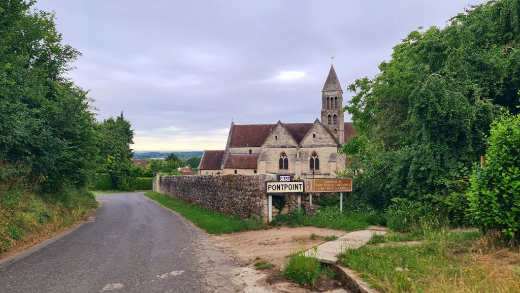 Een rustige weg leidt naar de kerk in Pontpoint, omringd door groene bomen en een grauwe lucht.