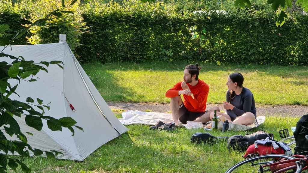 Twee jonge mensen zitten op een kleed voor een tent, genietend van een ontspannen moment op een camping. Ze hebben eten en drinken bij zich en bevinden zich in een groene omgeving met gras en struiken.