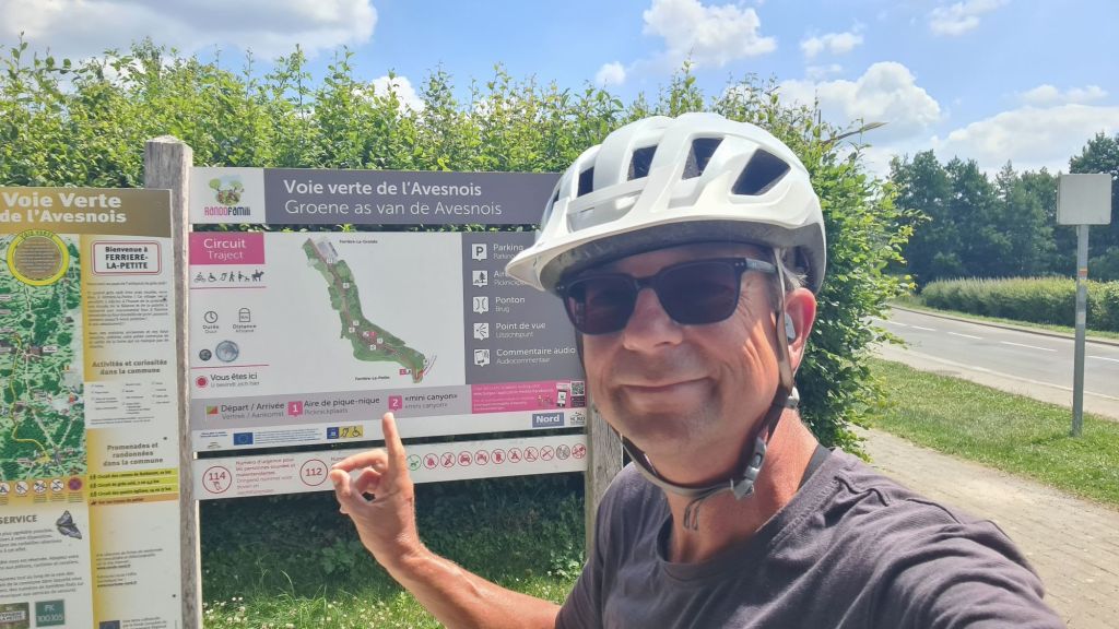 Een fietser staat bij een informatiebord van de Voie Verte de l'Avesnois, met een groene routekaart en instructies. De fietser draagt een helm en een zonnebril, en wijst naar het bord. Op de achtergrond zijn bomen en een blauwe lucht zichtbaar.
