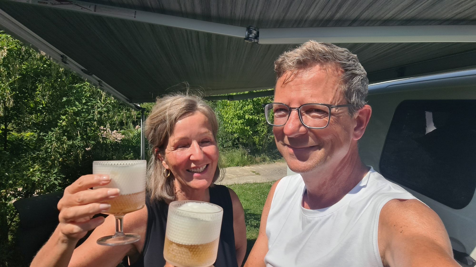 Twee mensen, een man en een vrouw, proosten met glazen gevuld met een bruisend drankje, terwijl ze samen poseren op een zonnige dag in de natuur.