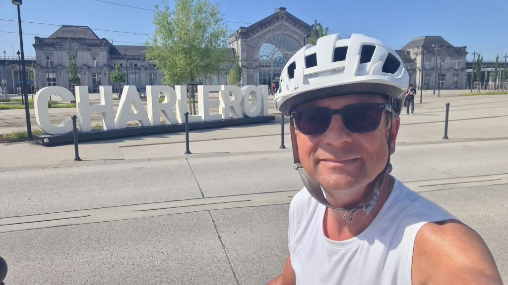 Een man op een fiets voor een groot bord met de tekst 'CHARLEROI' in een zonnige omgeving.