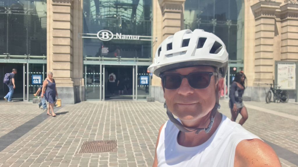 A person wearing a white helmet and sunglasses stands in front of the Namur train station, smiling. The entrance features a glass facade with a sign that reads 'B-Namur,' while people walk in and out of the station.