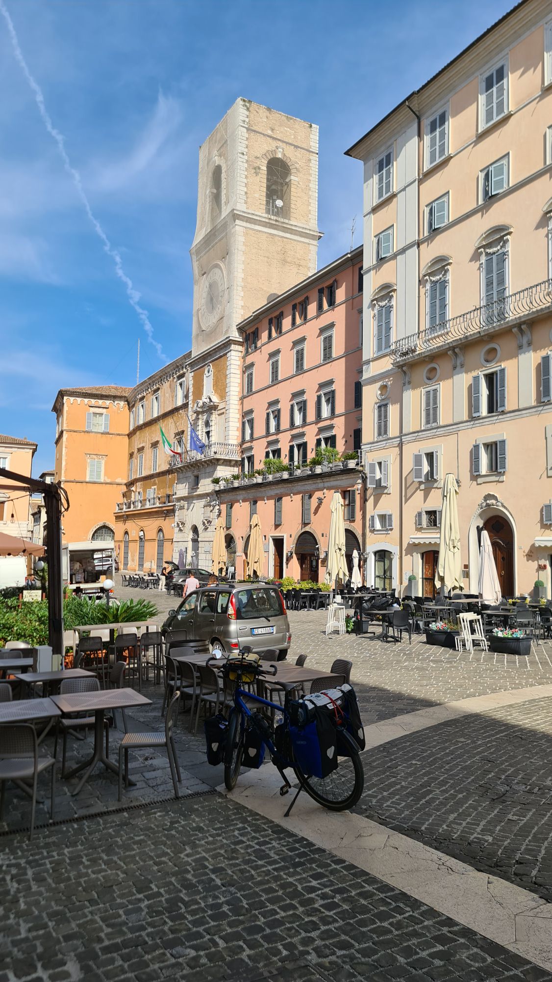 Een levendig plein in Italië met een historische kloktoren, kleurrijke gebouwen en een geparkeerde fiets met bagage.
