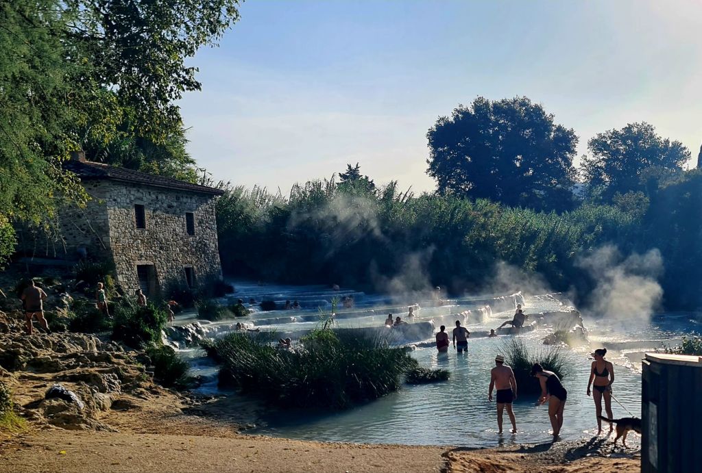 Een warmwaterbron in Saturnia met dampende thermen, omringd door groen. Mensen genieten van het warme water terwijl een stenen gebouw op de achtergrond zichtbaar is.