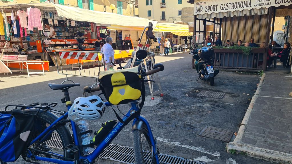 Een blauwe fiets met een helm en een fles water, naast een marktstraat vol kramen en mensen in een Italiaanse stad.