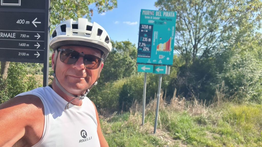 A cyclist taking a selfie in front of a sign indicating the 'Muro del Pirata', showing elevation and route details amidst greenery.