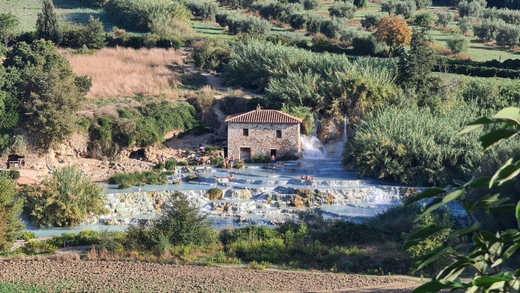 A picturesque view of natural hot springs with a stone building in the background, surrounded by greenery and people enjoying the warm waters.