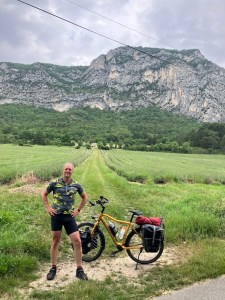 A person stands next to a yellow bicycle on a gravel path with green fields and a mountain range in the background.