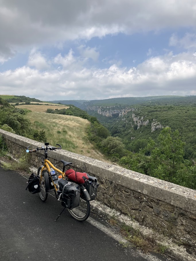 A yellow bicycle with saddlebags is parked next to a stone wall, overlooking a green valley with cliffs under a cloudy sky.