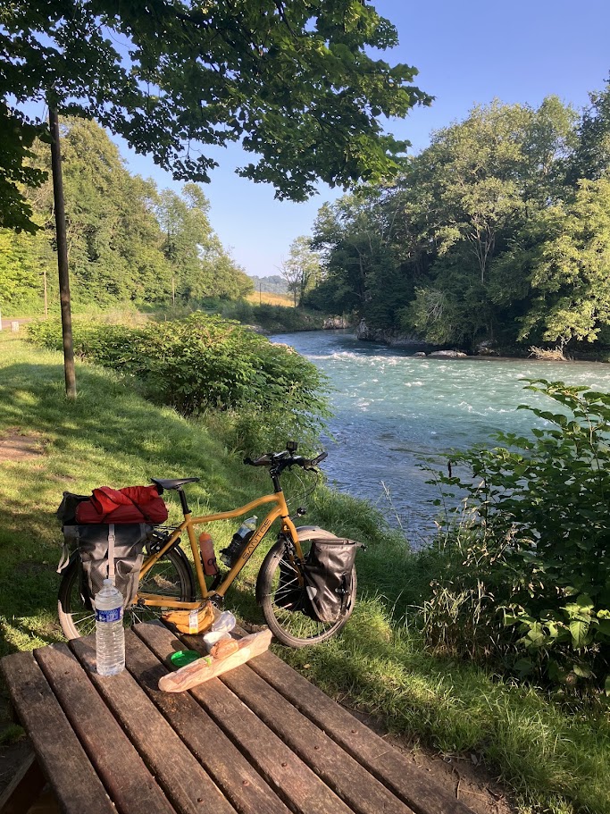 A bicycle with panniers parked by a peaceful river surrounded by lush greenery, with a wooden table and refreshments visible in the foreground.