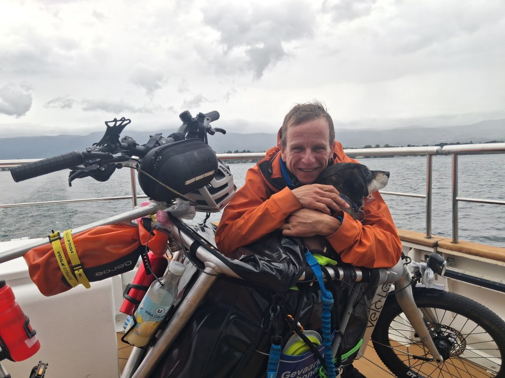 Fietsen met hond: Eus & Amma, een laatste grote reis: Een man in een oranje jas knuffelt een hond op een boot, met een fiets op de achtergrond en een meer en bergen in de verte.