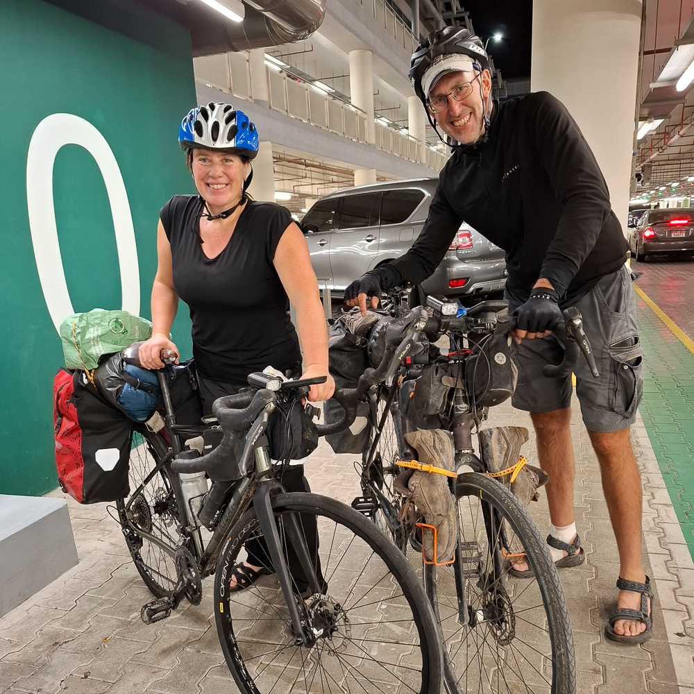 Podcast Van Vlaanderen naar de Gofl. Kristof en Roos staan naast hun fietsen in een parkeergarage, beiden in fietskleding en helmen, met verschillende tassen en uitrusting op hun fietsen.