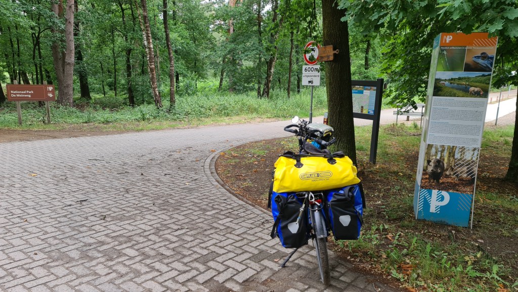 A bicycle with yellow panniers is parked near the entrance of National Park De Meinweg, with signage and a path leading into a lush green forest.