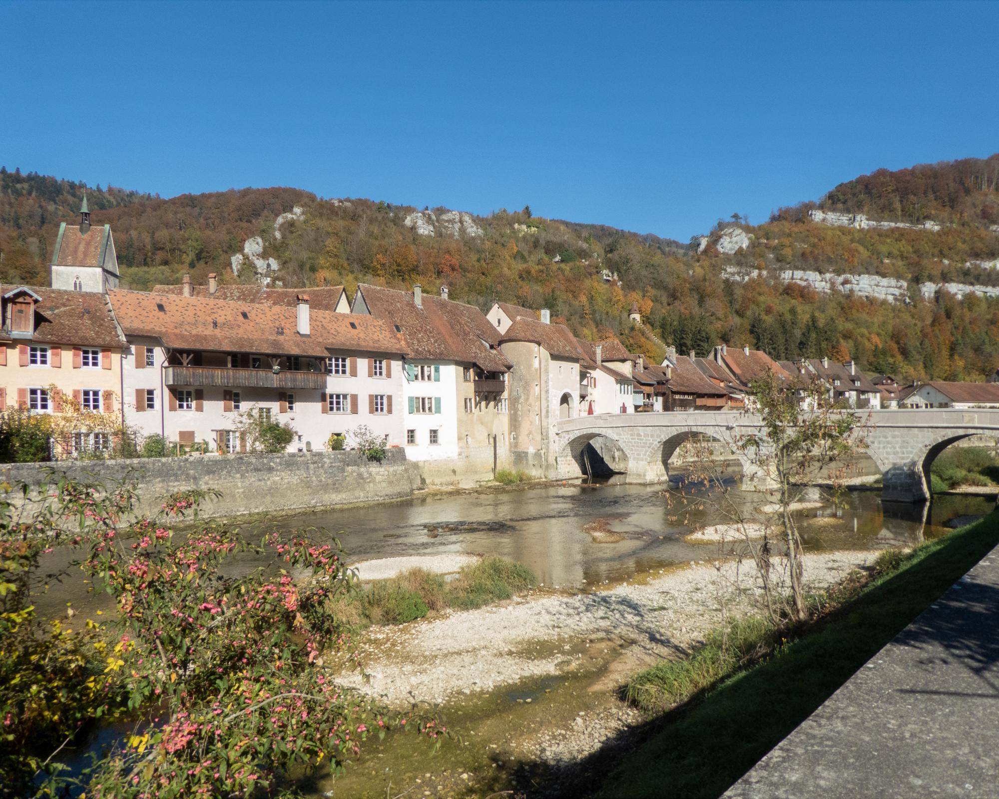 Stone bridge over a shallow river with old houses and a church tower against forested hills under a clear blue sky