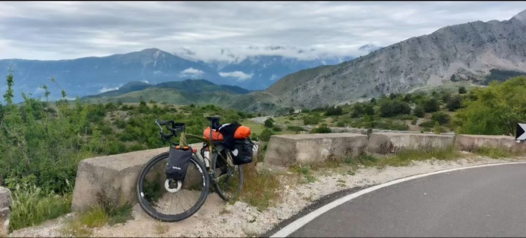 Fietsreis Albanië Rikkie Dijkhuizen.
Een fiets met bagage staat naast een weg met uitzicht op bergen en een groene vallei onder een bewolkte lucht.