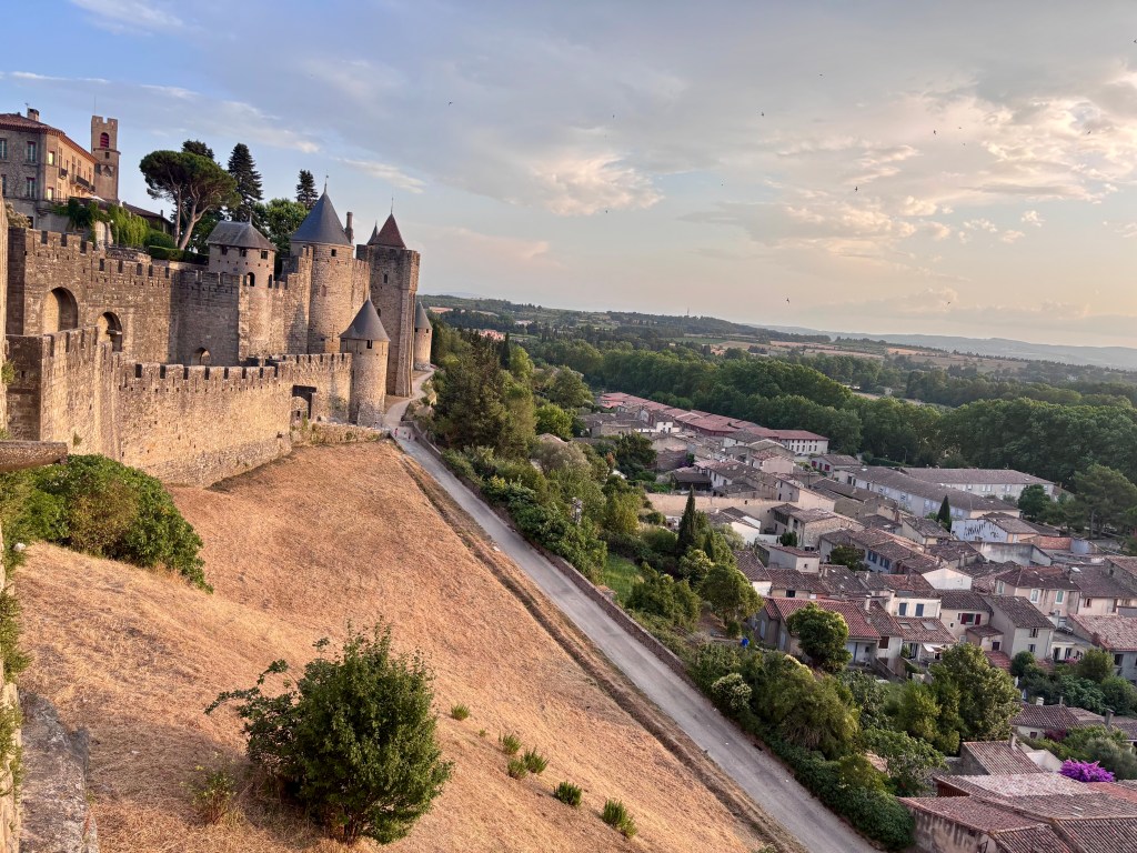 100 cols een groot verlangen. Uitzicht op een middeleeuwse vesting met torens en muren, omringd door groene heuvels en een dorp in de vallei.