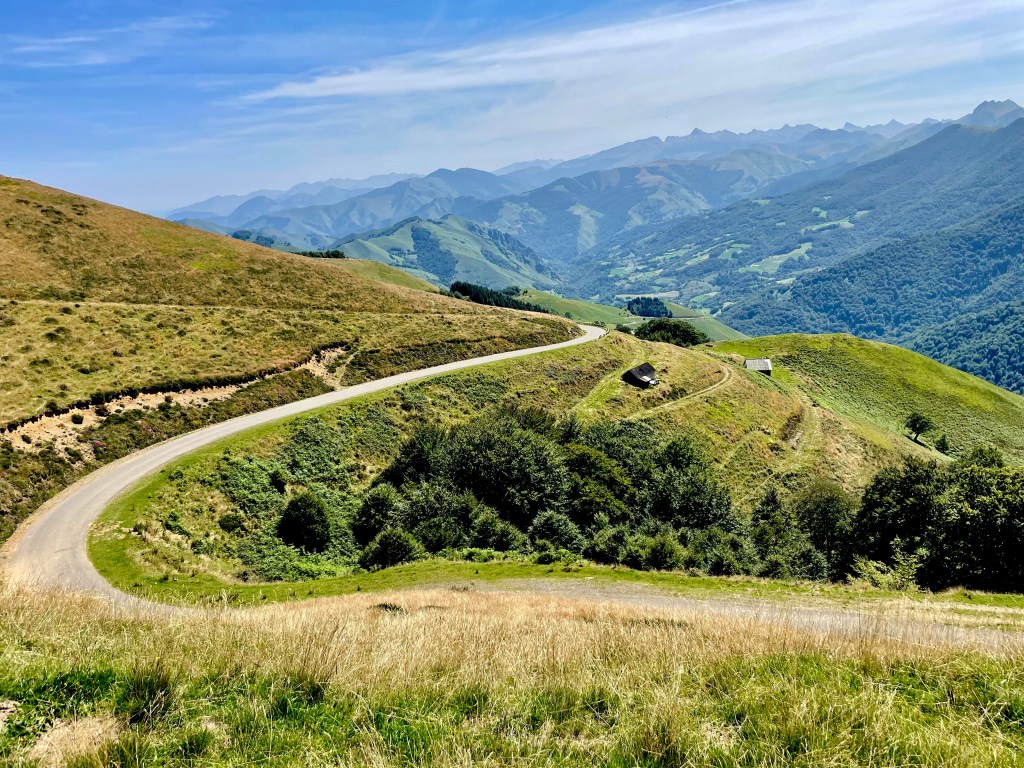 100 cols een groot verlangen. Landschap met een kronkelige weg die door groene heuvels slingert, omringd door bergen en een heldere lucht.