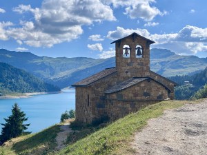 100 cols een groot verlangen. Een kerk met 2 klokken met op de achtergrond een blauw meer tussen de bergen die groen zijn begroeid.